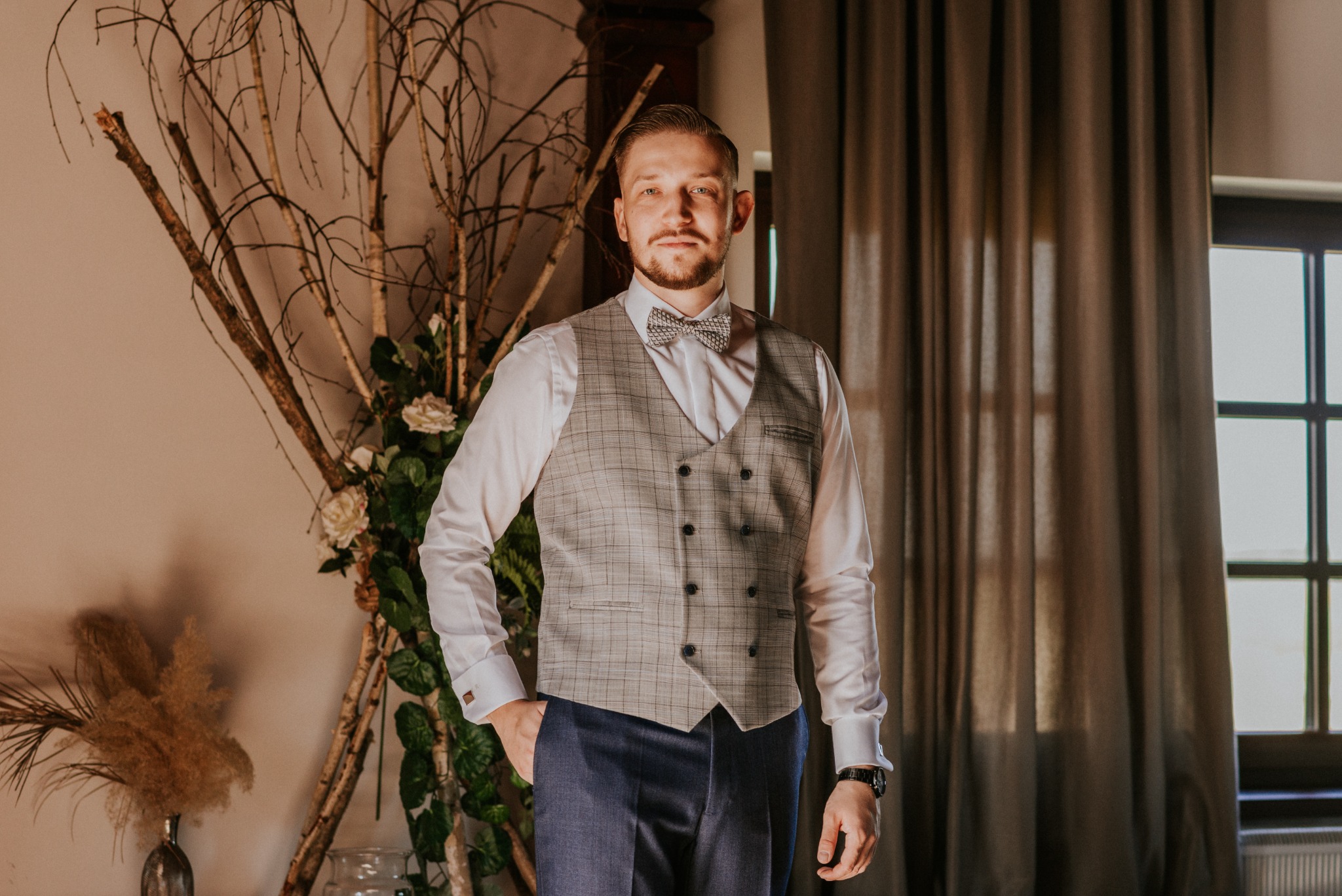 Elegancki pan młody w kraciastej kamizelce i muszce, pozujący na tle dekoracji z gałęzi i kwiatów, z ręką w kieszeni spodni.