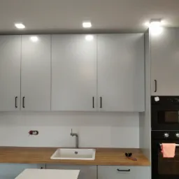 Szara zabudowa kuchenna z drewnianym blatem, widoczny zlew, piekarnik do zabudowy i narzędzia na blacie, sugerujące prace wykończeniowe.