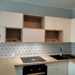 Nowoczesna kuchnia z białymi szafkami, drewnianymi blatami i geometryczną mozaiką na ścianie, widok na płytę indukcyjną i zlewozmywak, oświetlenie sufitowe z metalowymi kloszami.