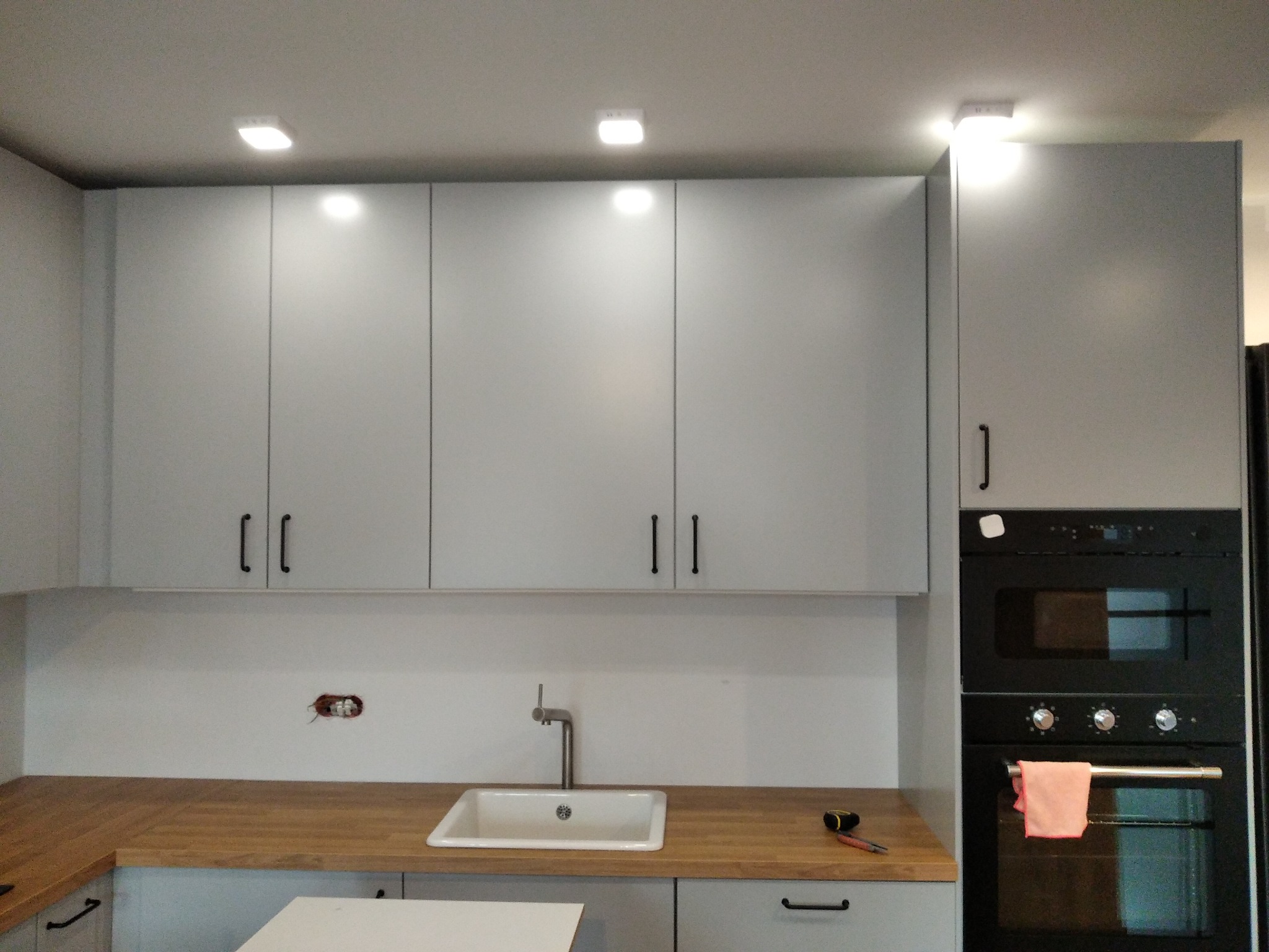 Szara zabudowa kuchenna z drewnianym blatem, widoczny zlew, piekarnik do zabudowy i narzędzia na blacie, sugerujące prace wykończeniowe.