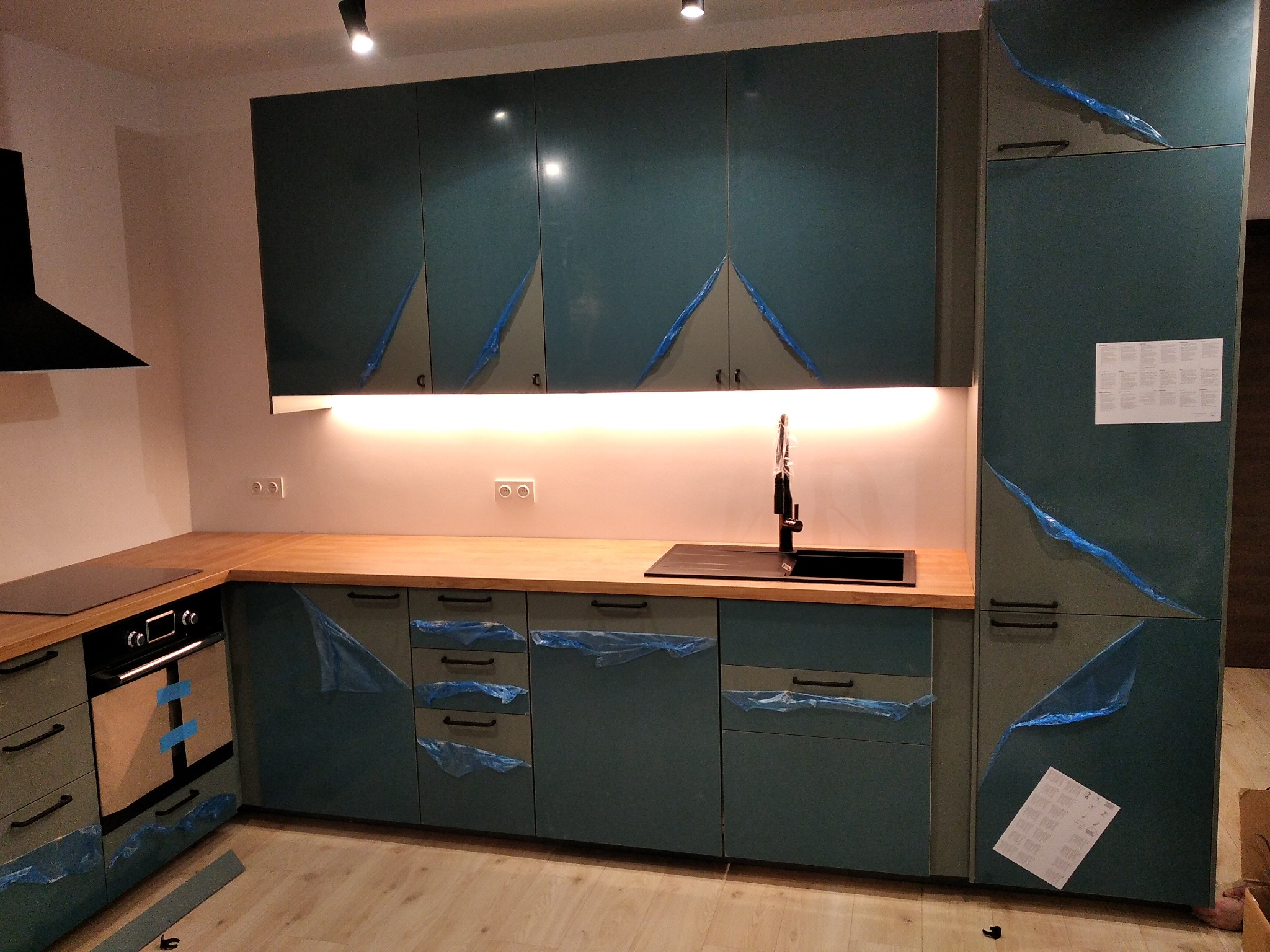 Nowoczesna kuchnia z zielonymi frontami, drewnianym blatem i czarnym zlewem, jeszcze zabezpieczona folią ochronną po montażu.