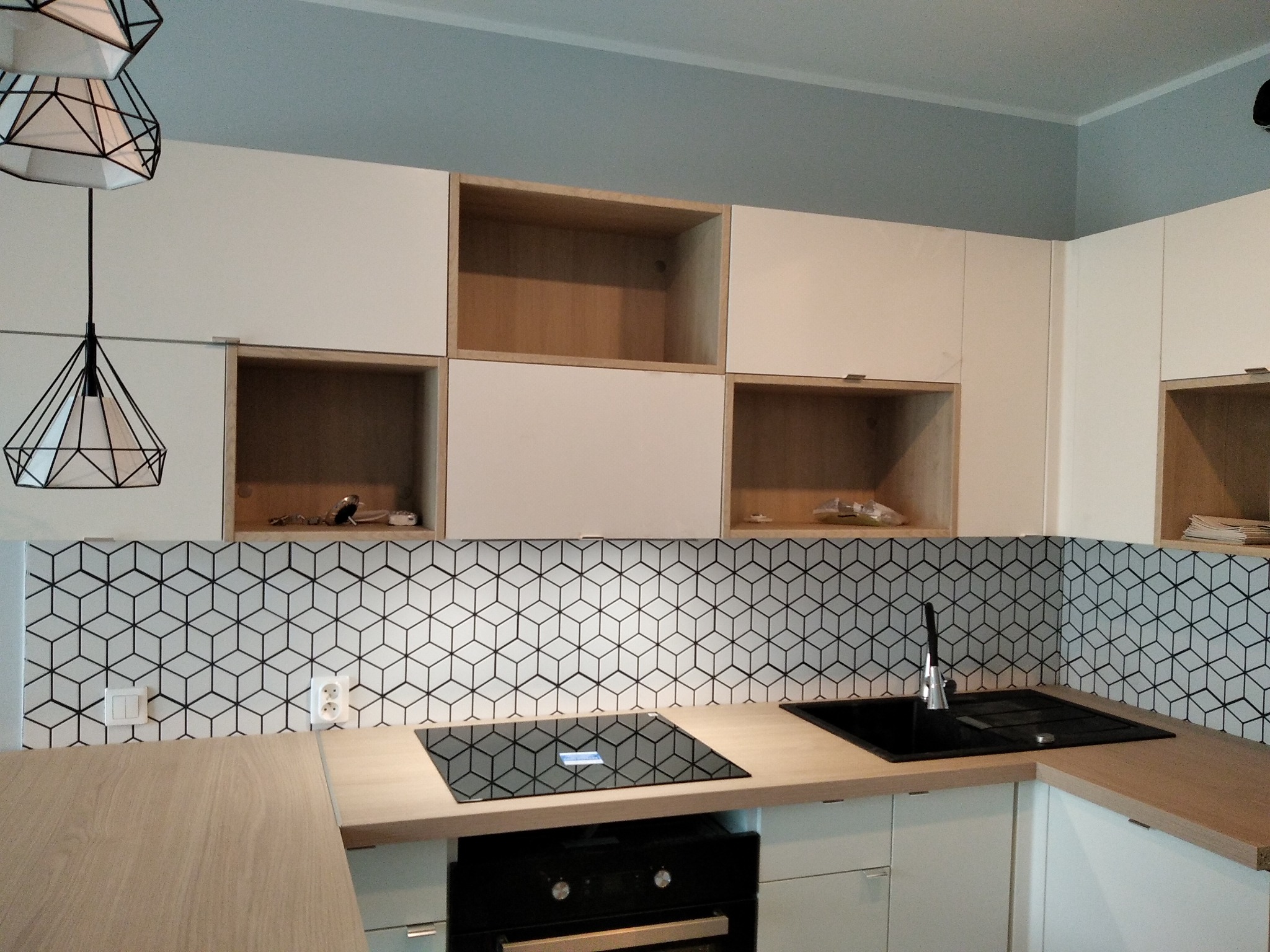 Nowoczesna kuchnia z białymi szafkami, drewnianymi blatami i geometryczną mozaiką na ścianie, widok na płytę indukcyjną i zlewozmywak, oświetlenie sufitowe z metalowymi kloszami.