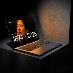 Laptop z wyświetloną stroną internetową prezentującą portret kobiety i daty 1874-2025, umieszczony na betonowej powierzchni w minimalistycznym, ciemnym otoczeniu.