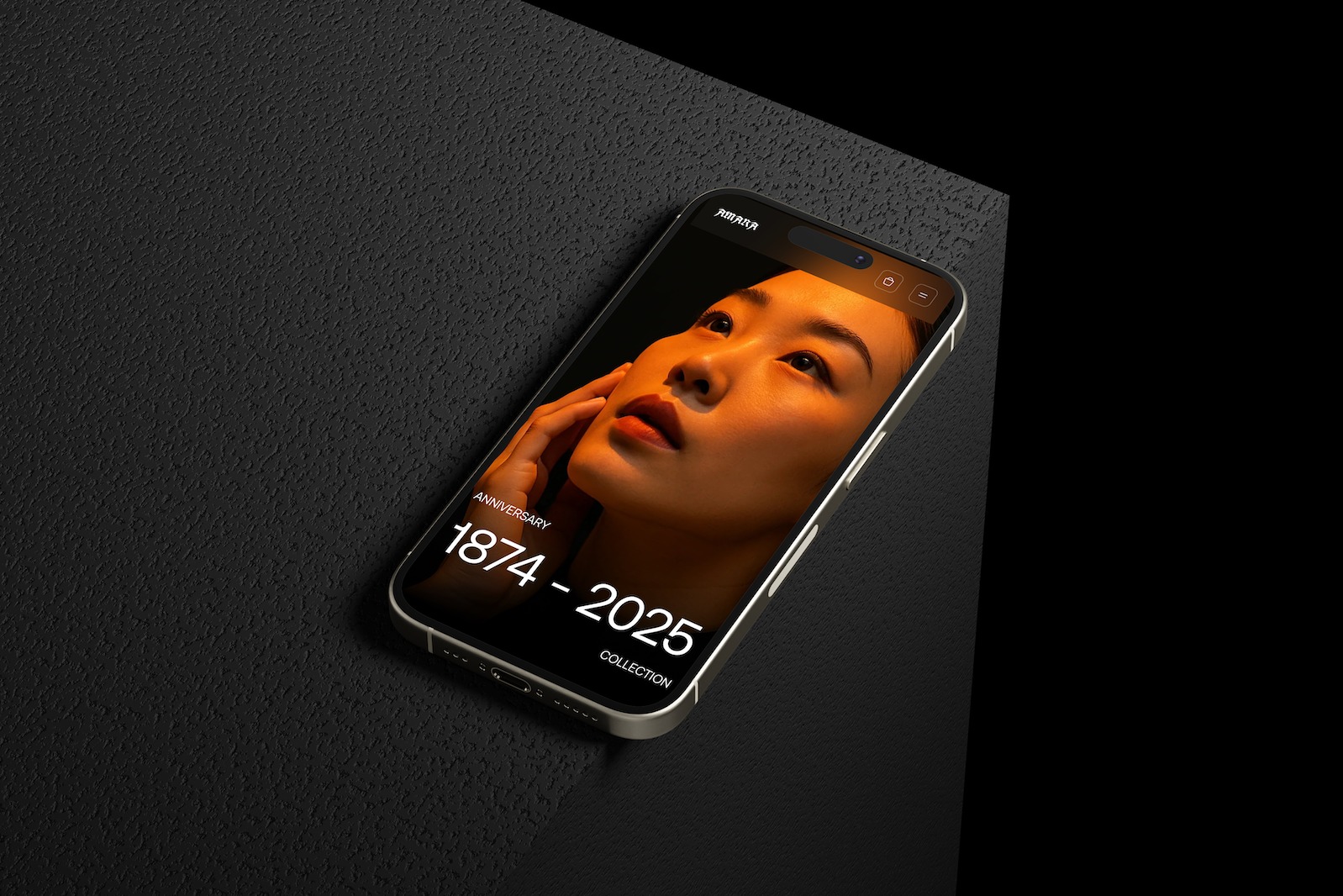 Smartfon z wyświetlonym portretem kobiety w ciepłych barwach i napisem 'Anniversary 1874-2025 Collection', leżący na teksturowanym, ciemnym tle.