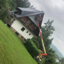 Michał Szwed G - Roof - Praca na wysokości: operator w podnośniku koszowym przy dachu domu z ciemną dachówką, otoczonego zielenią. Widok na dom z trawnikiem i drzewami w tle.