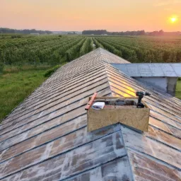 Metalowy dach w trakcie naprawy, z widocznym drewnianym obramowaniem komina, poziomnicą, notatnikiem, wiertarką akumulatorową i rozległym sadem w tle o zachodzie słońca.