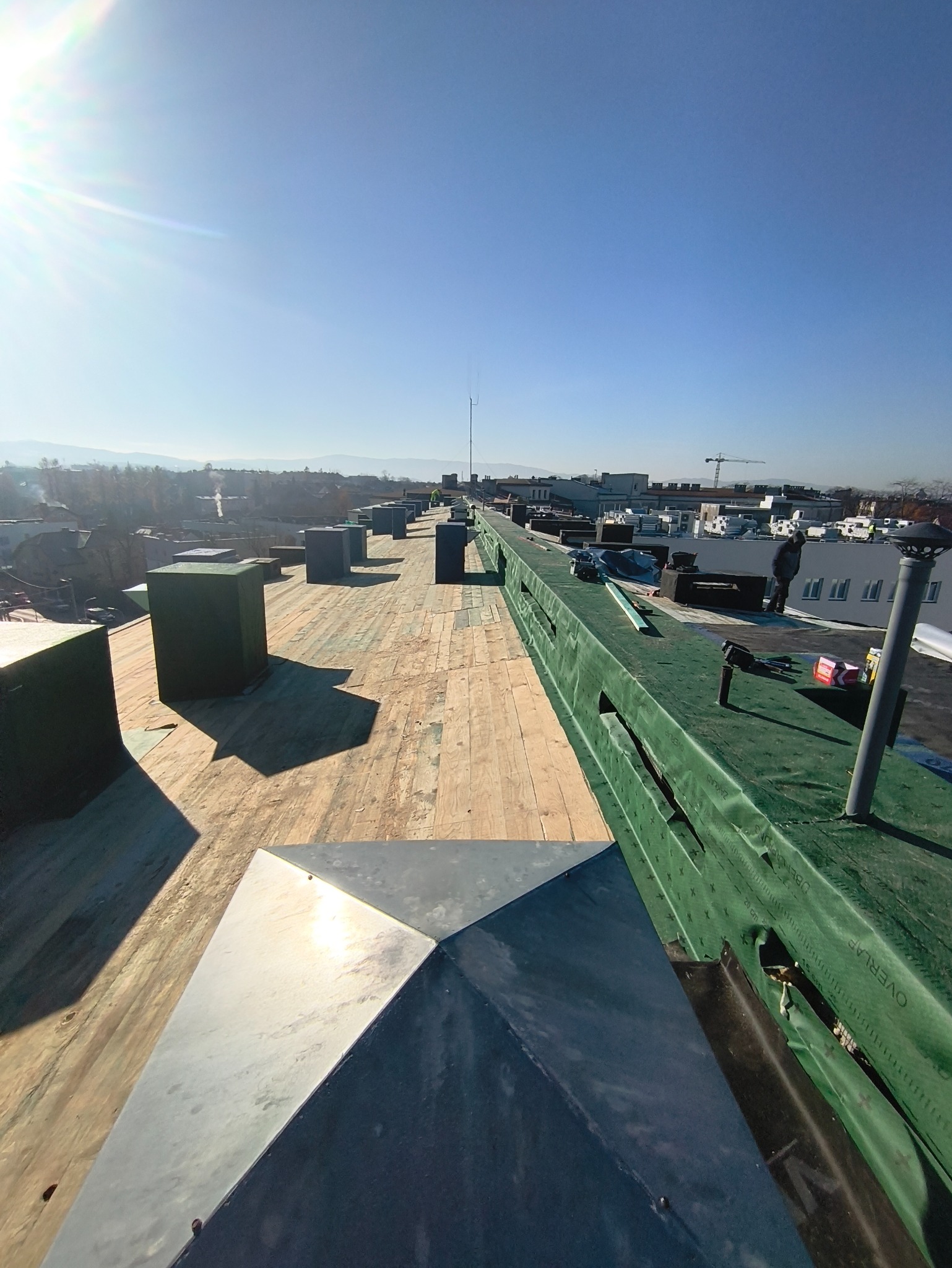 Dach budynku w trakcie krycia, widoczne drewniane deski, elementy wentylacyjne i zabezpieczenia. W tle zabudowa miejska i błękitne niebo.