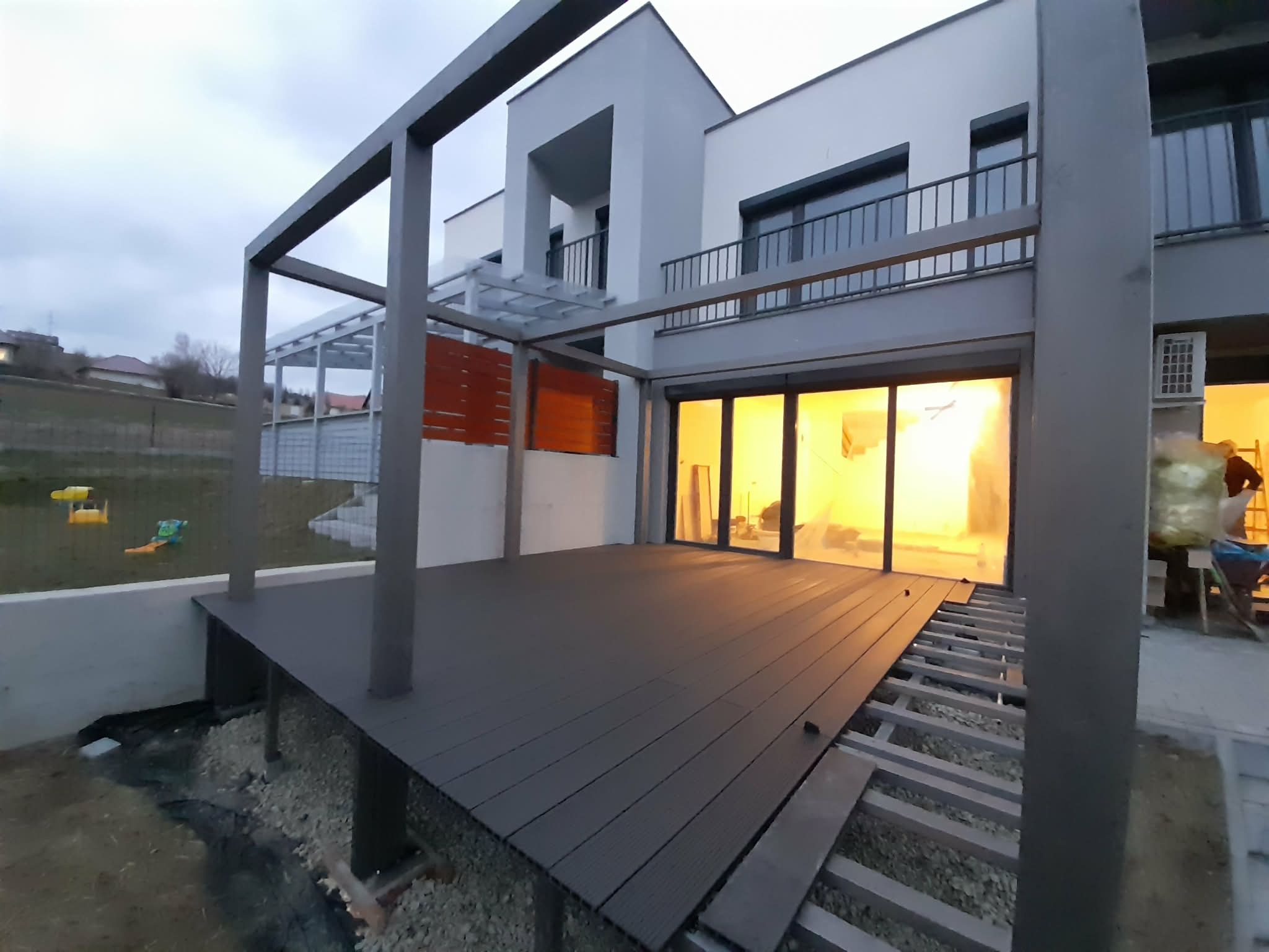 Nowoczesny taras kompozytowy w trakcie budowy, z widoczną konstrukcją nośną i częściowo ułożonymi deskami, na tle domu z dużymi oknami.