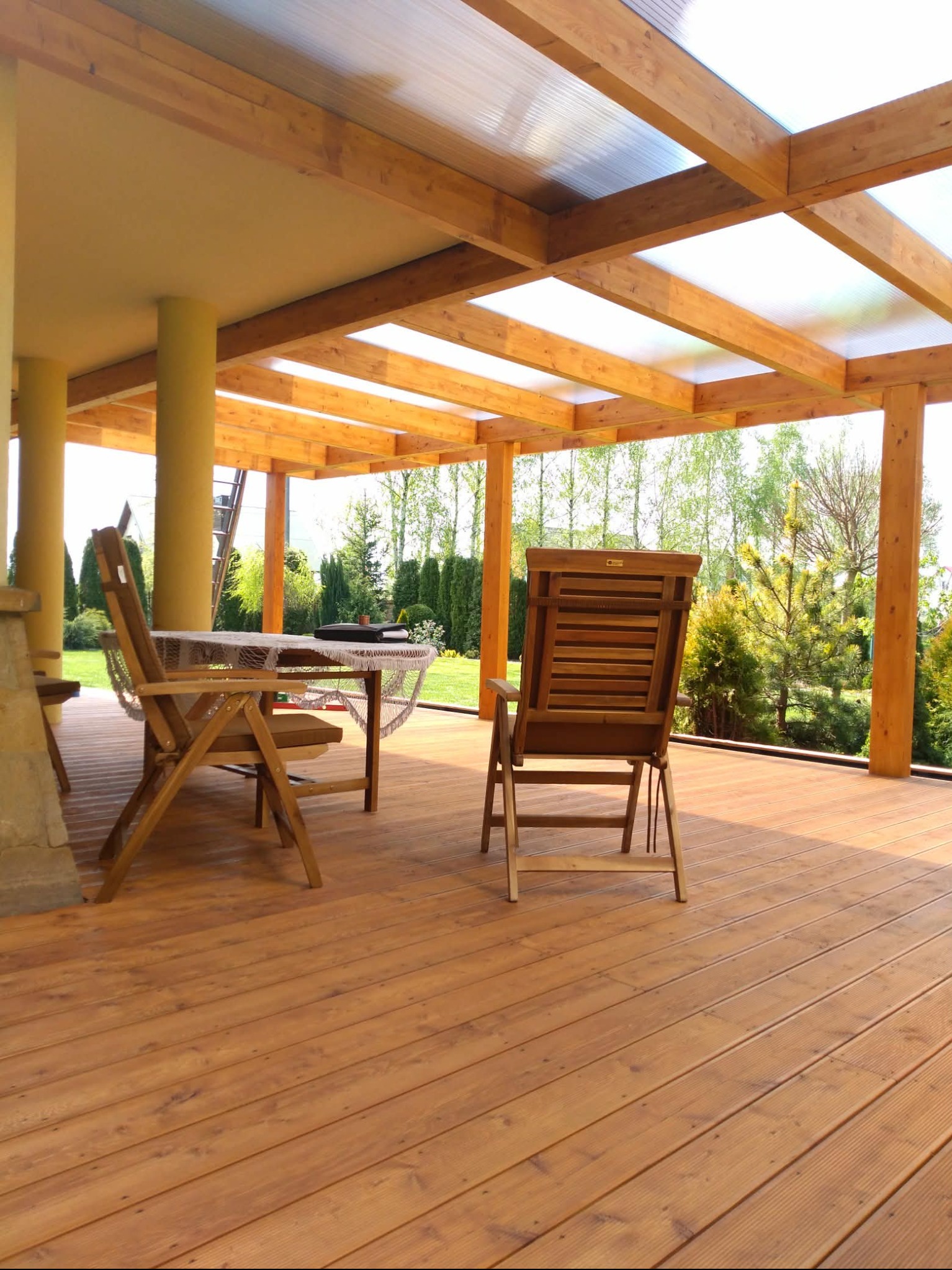 Zadaszony taras kompozytowy z drewnianą konstrukcją, meblami ogrodowymi i widokiem na zieleń. Jasne, naturalne światło podkreśla ciepły odcień drewna.