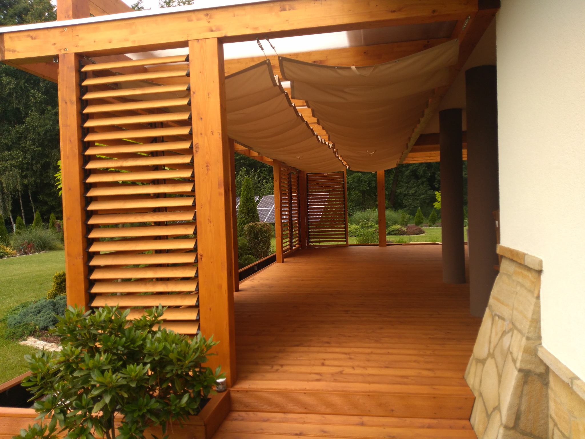 Drewniana pergola z żaluzjami i płóciennym zadaszeniem, widok na taras i ogród. Elementy konstrukcyjne w ciepłym odcieniu drewna, zieleń w tle.