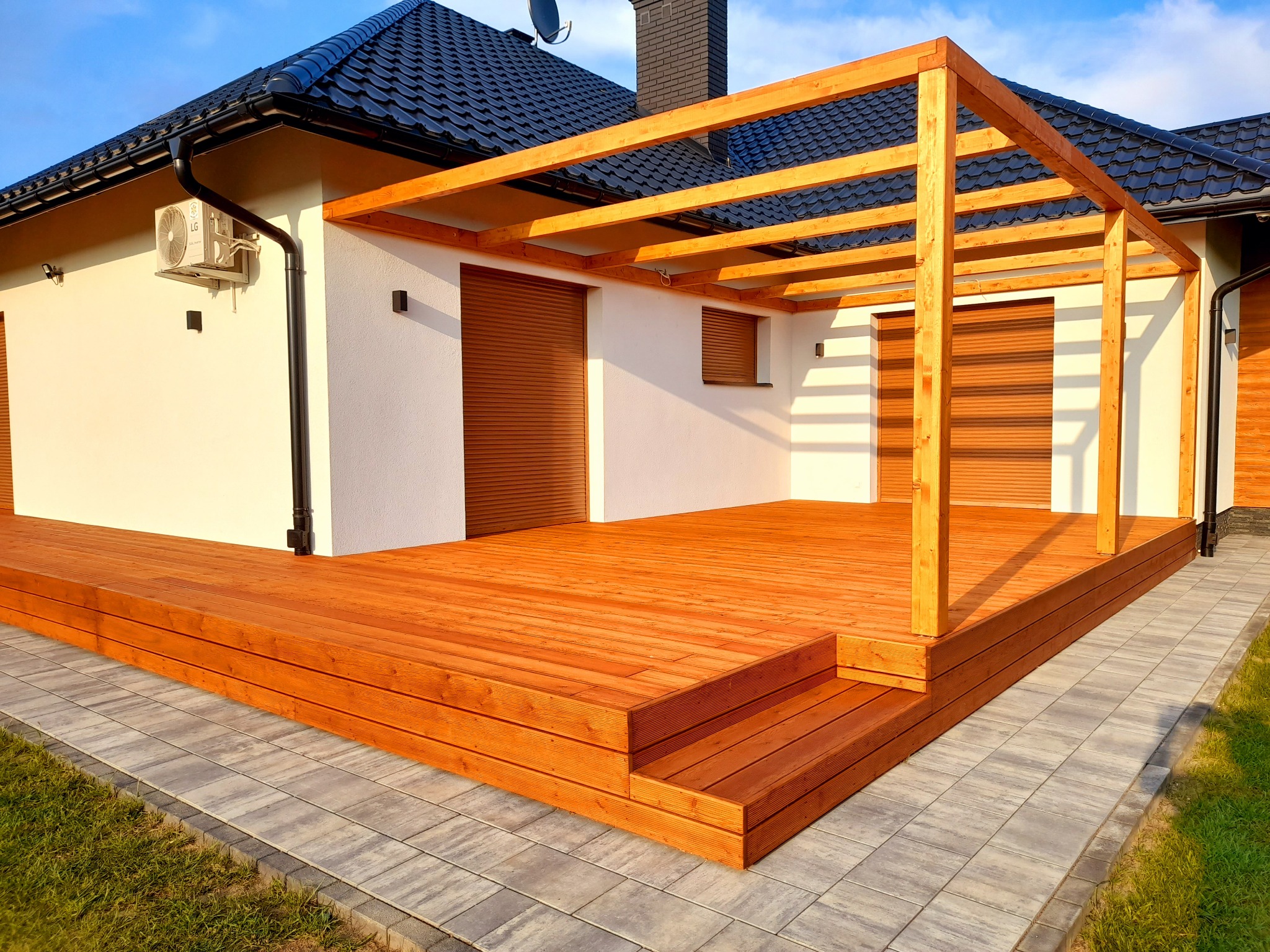 Nowoczesny taras kompozytowy z drewnianą pergolą, schodami i kostką brukową, przylegający do białego budynku z czarnym dachem w słoneczny dzień.