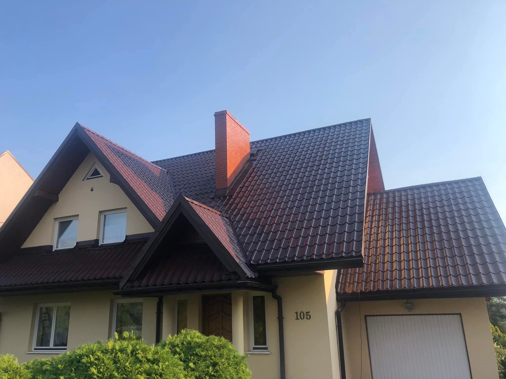 Ciemnobrązowy dach z blachodachówki na beżowym domu z ceglanym kominem, numer domu 105, widok z dołu w słoneczny dzień.