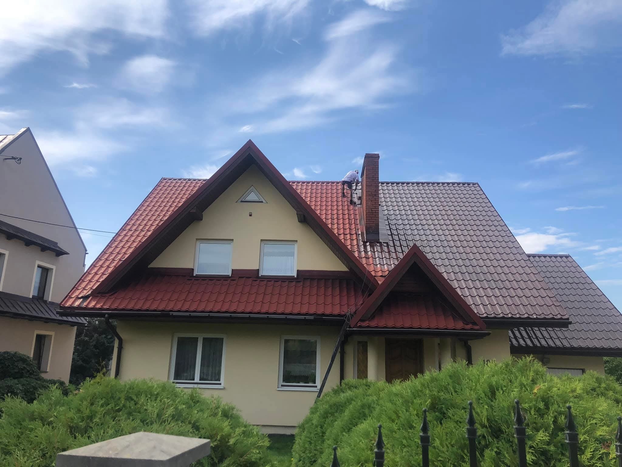 Częściowo pomalowany dach z czerwonej dachówki, widoczny pracownik na dachu, drabina oparta o ścianę domu, błękitne niebo z chmurami w tle.