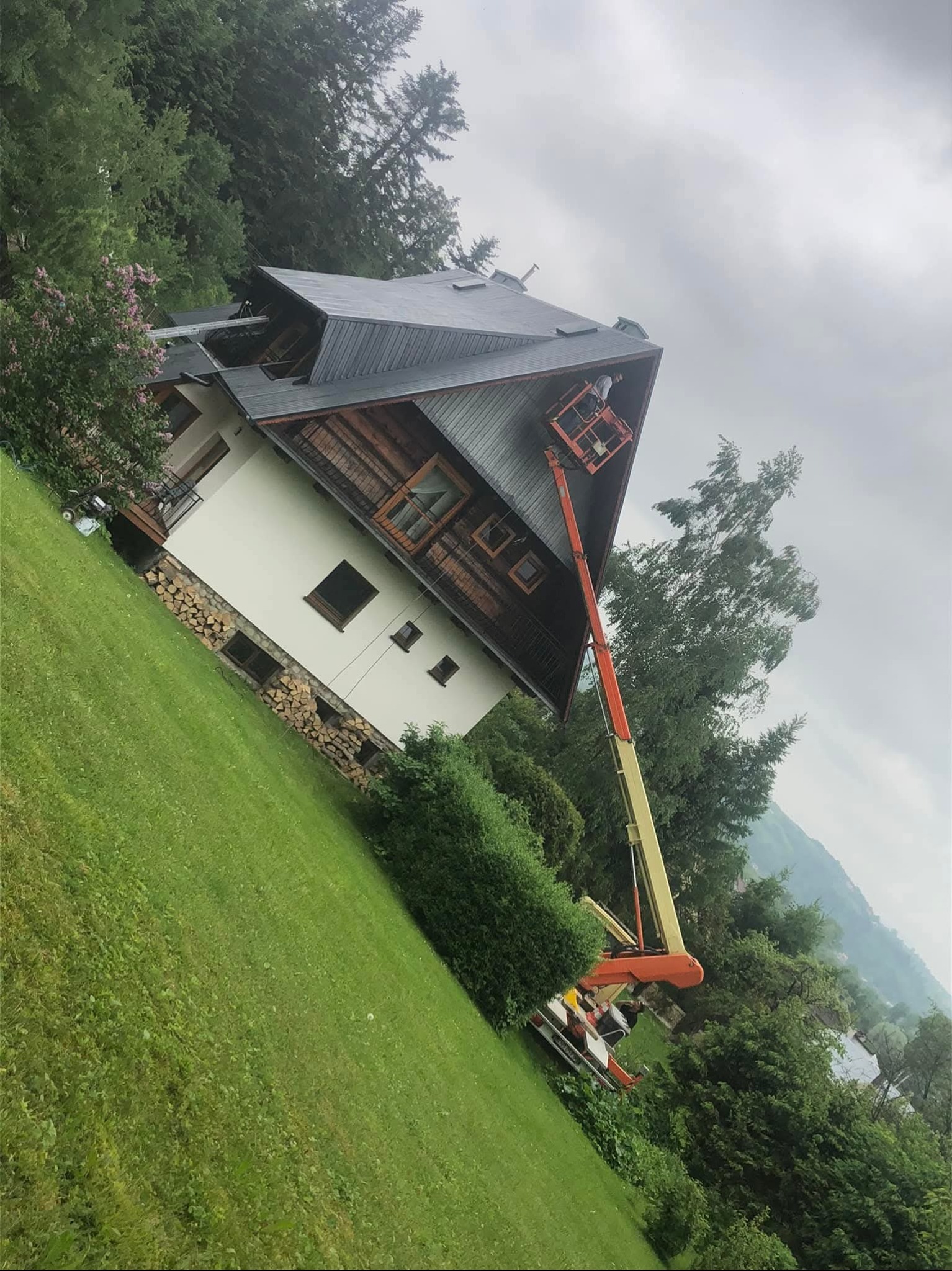 Praca na wysokości: operator w podnośniku koszowym przy dachu domu z ciemną dachówką, otoczonego zielenią. Widok na dom z trawnikiem i drzewami w tle.