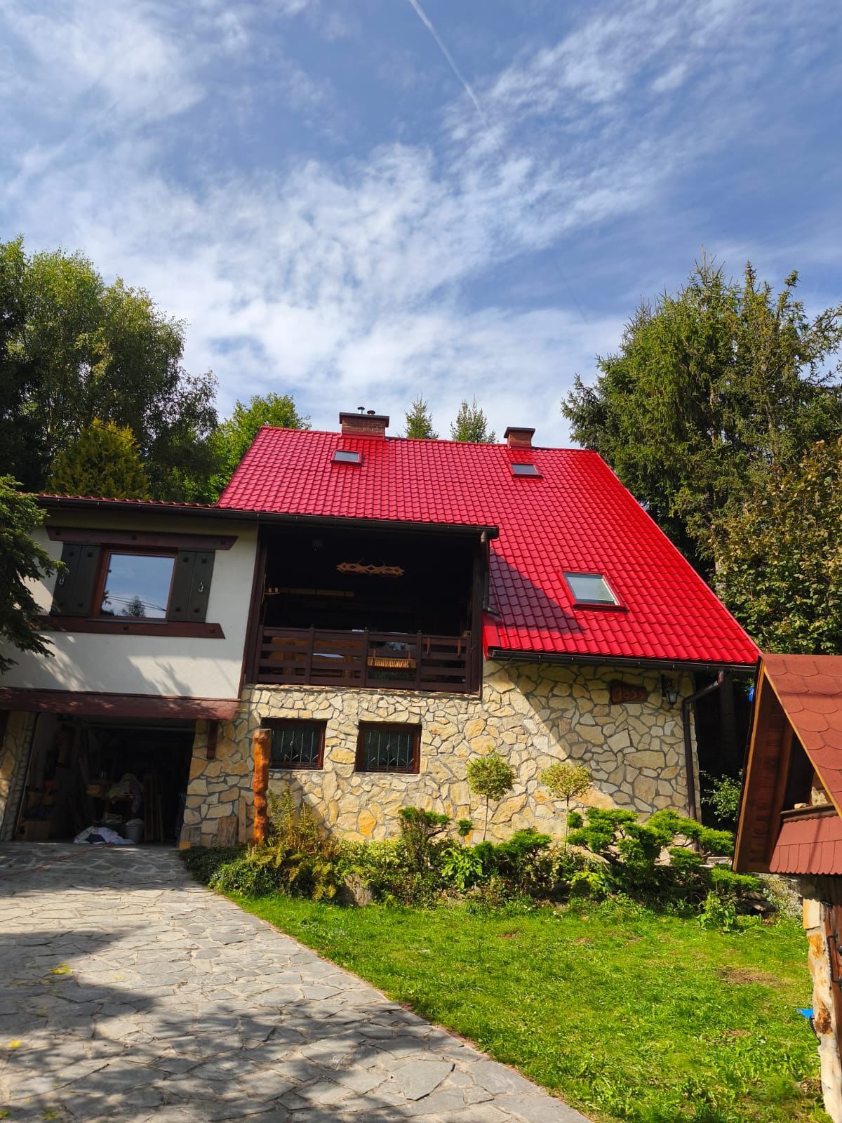 Dom z kamienną elewacją i intensywnie czerwonym dachem z dachówki, otoczony zielenią. Widoczne okna dachowe i balkon z drewnianą balustradą. Słoneczny dzień.
