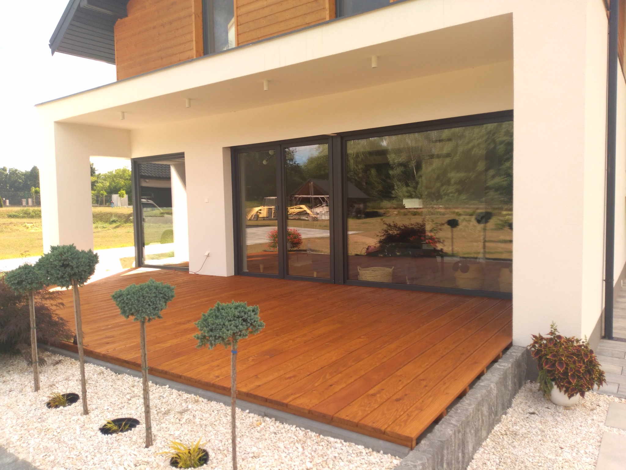 Nowoczesny taras drewniany z desek w ciepłym odcieniu brązu, otoczony białym żwirem i ozdobnymi drzewkami, przylegający do budynku z dużymi, przeszklonymi drzwiami.