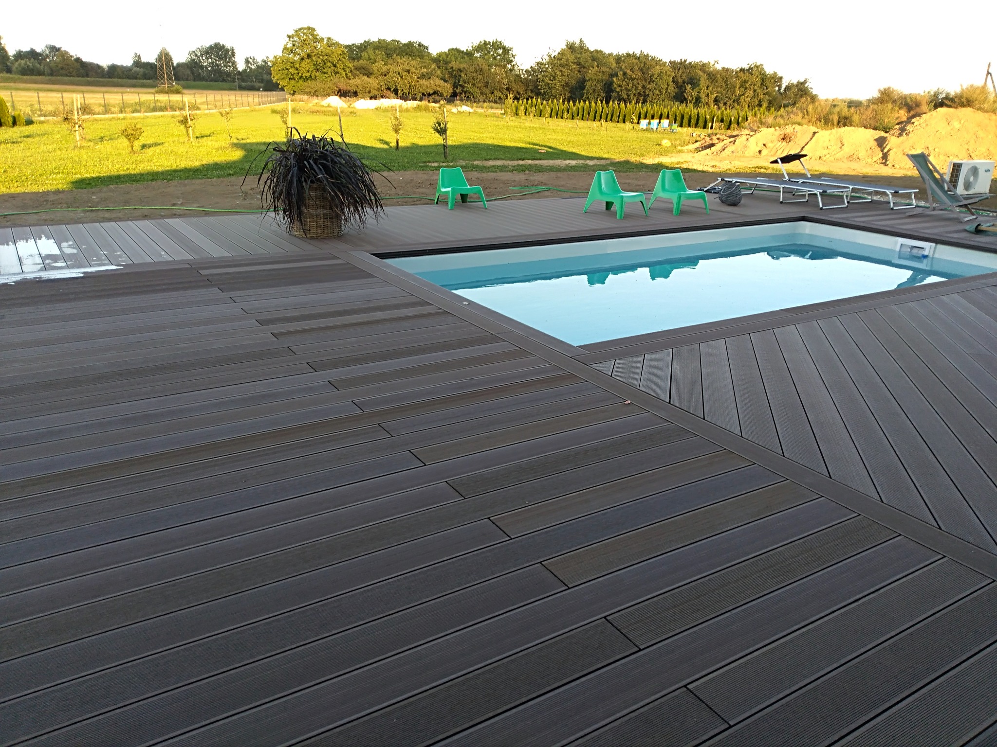 Nowoczesny taras kompozytowy w odcieniach szarości otacza basen. Widoczne zielone krzesła i leżaki. W tle trawnik i drzewa, tworzące relaksującą przestrzeń.