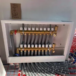 Rozdzielacz ogrzewania podłogowego w białej skrzynce, z podłączonymi rurami grzewczymi na folii izolacyjnej, obok kartka z odręcznymi notatkami o nazwach pomieszczeń.