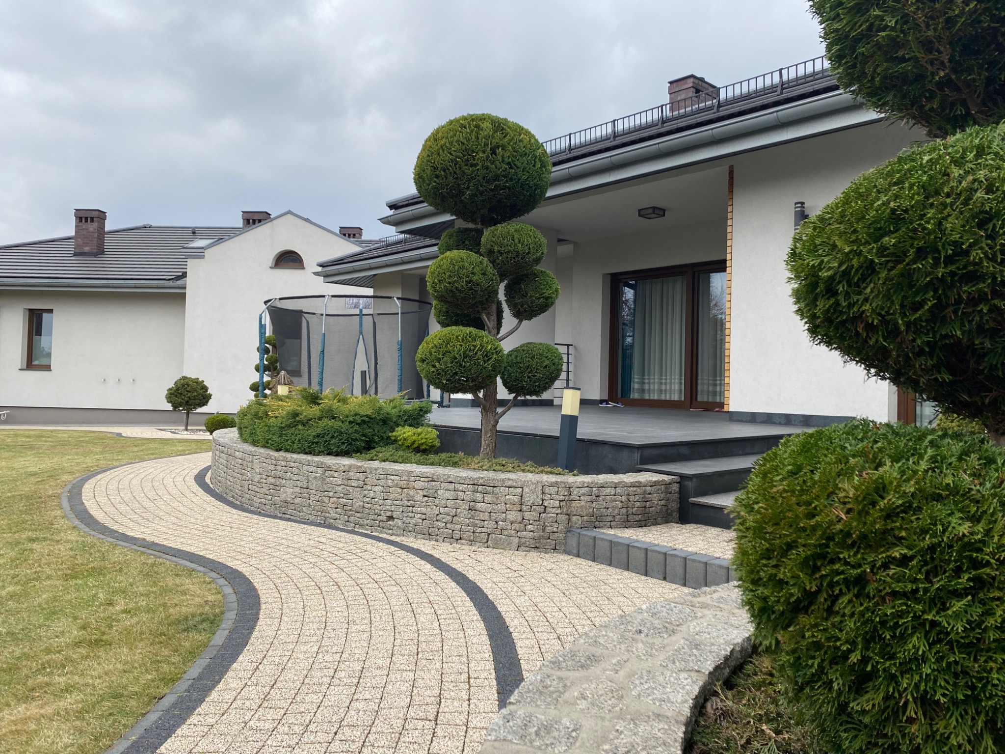 Kaskadowy ogród z krzewami formowanymi w kule, ścieżka z jasnej kostki brukowej z ciemnym obramowaniem prowadząca do domu z ciemnymi schodami i szarym dachem