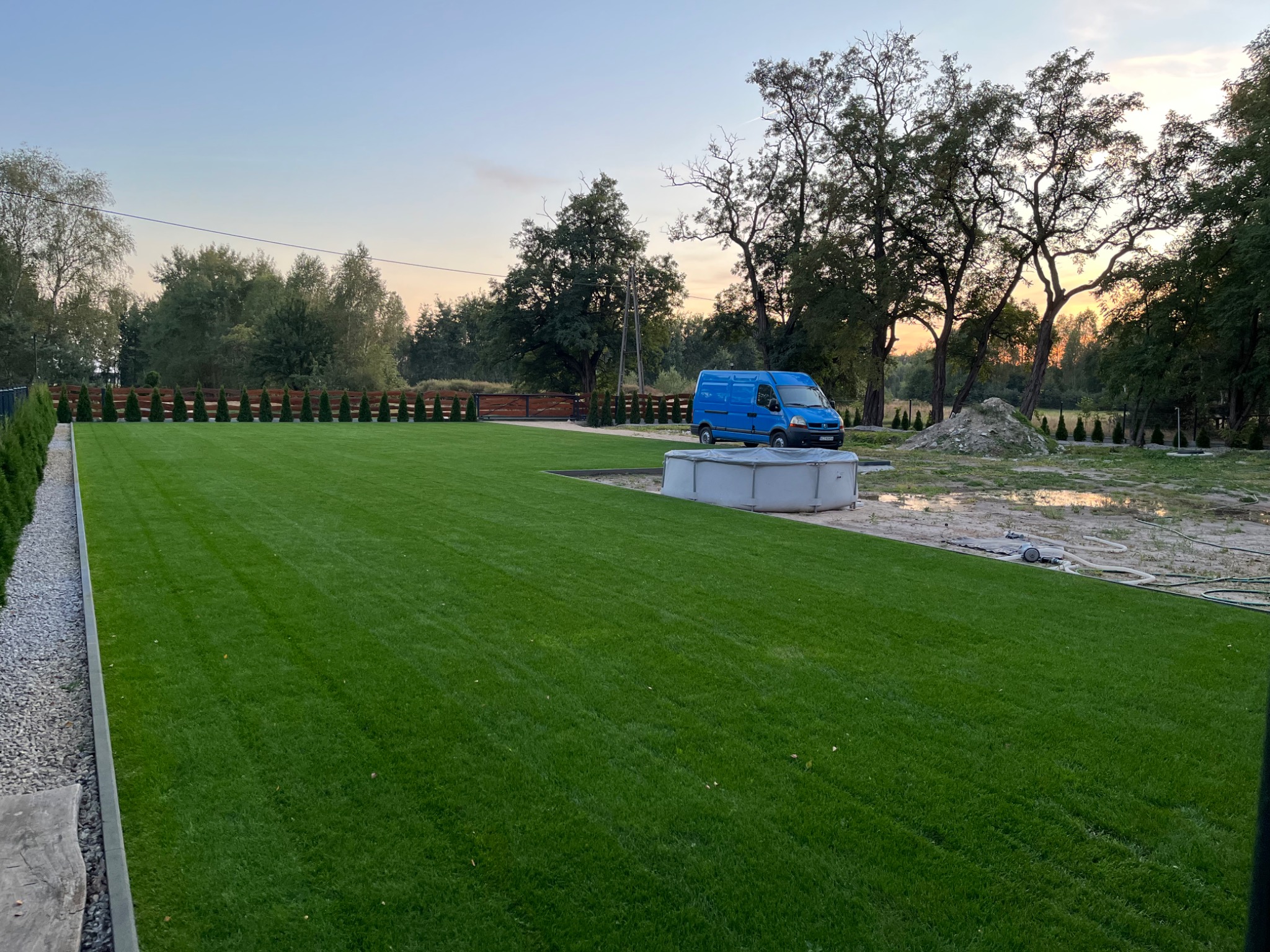 Nierówny teren po niwelacji z wygładzoną, intensywnie zieloną trawą, z boku zaparkowany niebieski bus firmy, w tle drzewa i zachodzące słońce.