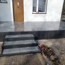 Fil-bruk - Wykończony podest wejściowy z szarej kostki, prowadzący do drzwi wejściowych domu, z trzema stopniami i rabatą z wrzosami z boku.