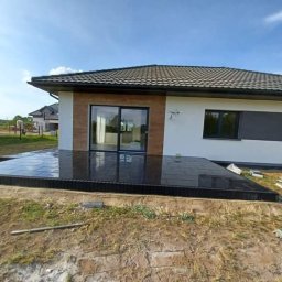 Fil-bruk - Nowoczesny taras z ciemnej, błyszczącej kostki przy białym domu z dużymi oknami przesuwnymi, widok na ogród w słoneczny dzień.