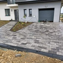Fil-bruk - Ułożona kostka brukowa w odcieniach szarości prowadząca do domu z garażem, widoczny czarny pies na kostce i świeżo zasadzone rośliny wzdłuż ścieżki.