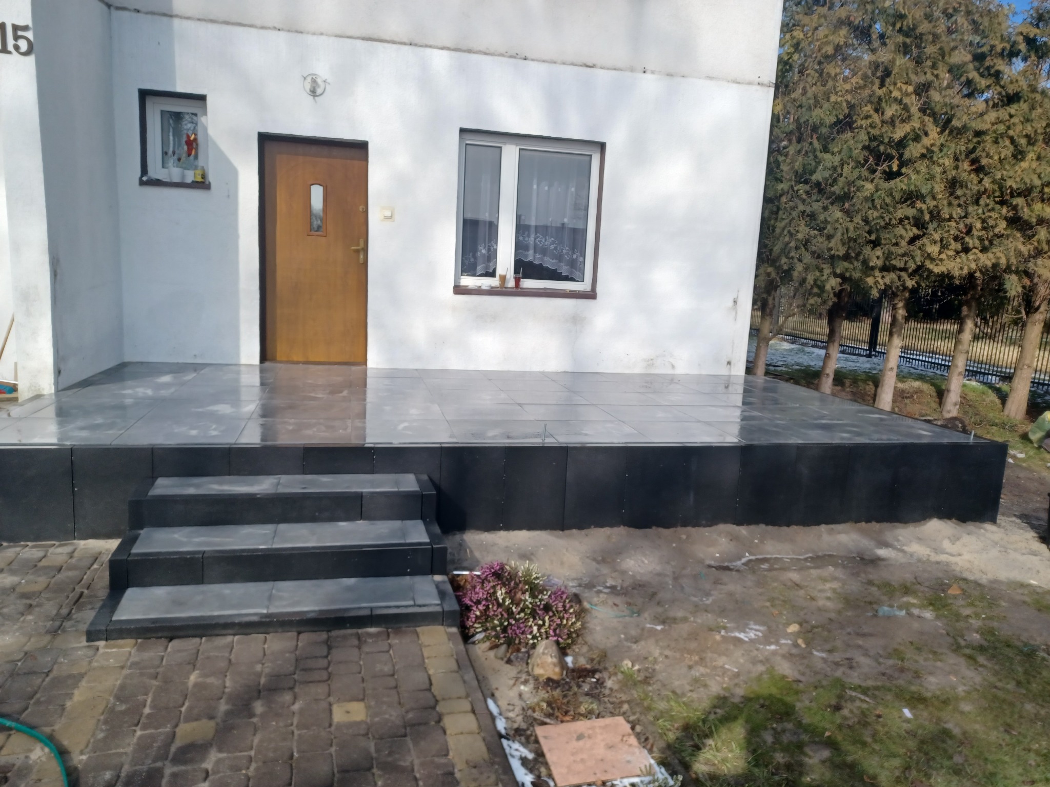 Wykończony taras z szarych płytek, z trzema stopniami z ciemnej kostki, prowadzącymi do drzwi wejściowych białego domu. Na pierwszym planie kostka brukowa i mały krzew. W tle widoczne drzewa.