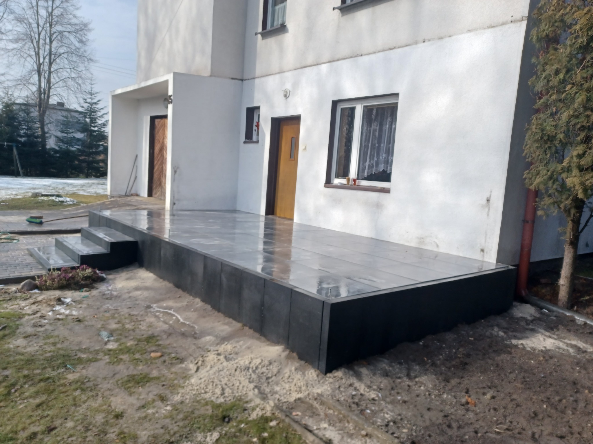 Nowoczesny taras z szarych płyt ułożonych przed białym domem z drewnianymi drzwiami, widoczne schody wejściowe i fragment ogrodu z drzewami.