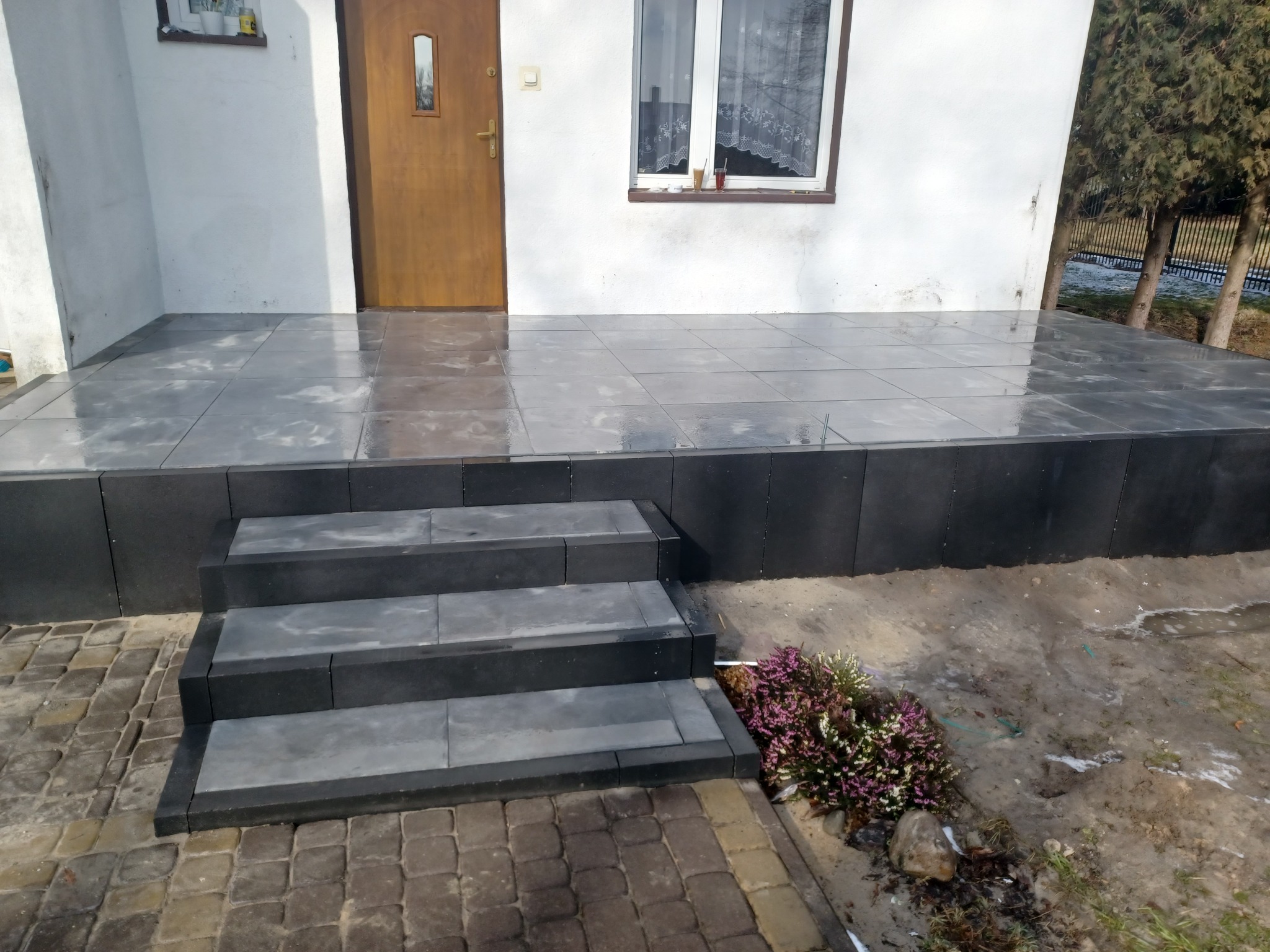 Wykończony podest wejściowy z szarej kostki, prowadzący do drzwi wejściowych domu, z trzema stopniami i rabatą z wrzosami z boku.