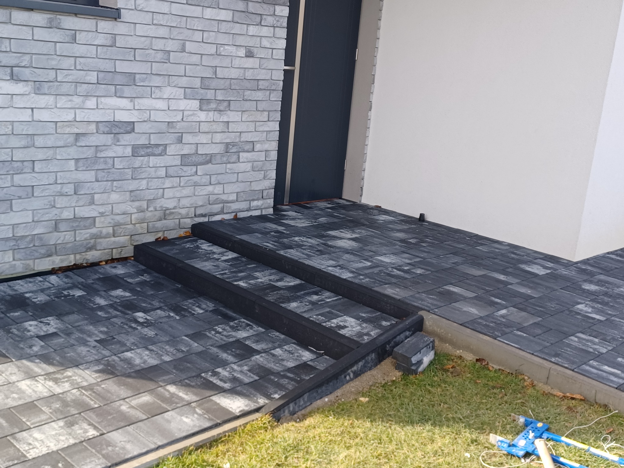 Ułożona szara kostka brukowa prowadząca do drzwi wejściowych, z widocznymi stopniami i narzędziami brukarskimi na trawniku.