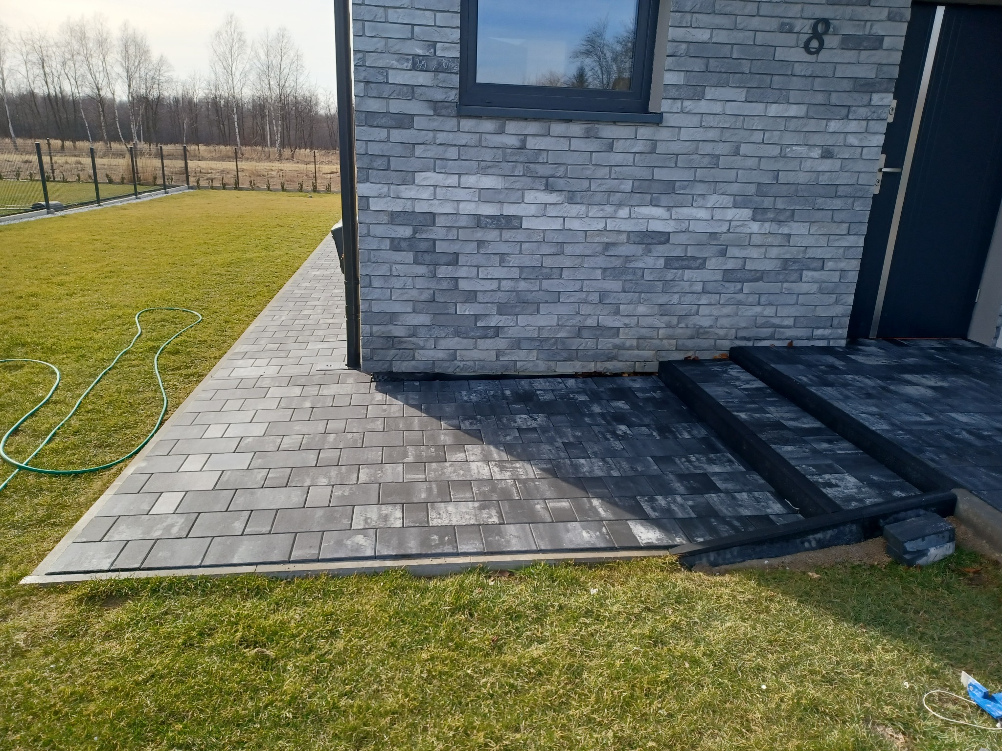 Ułożona ścieżka z szarej kostki brukowej prowadząca do domu z elewacją z szarej cegły, widoczny trawnik i fragment węża ogrodowego.