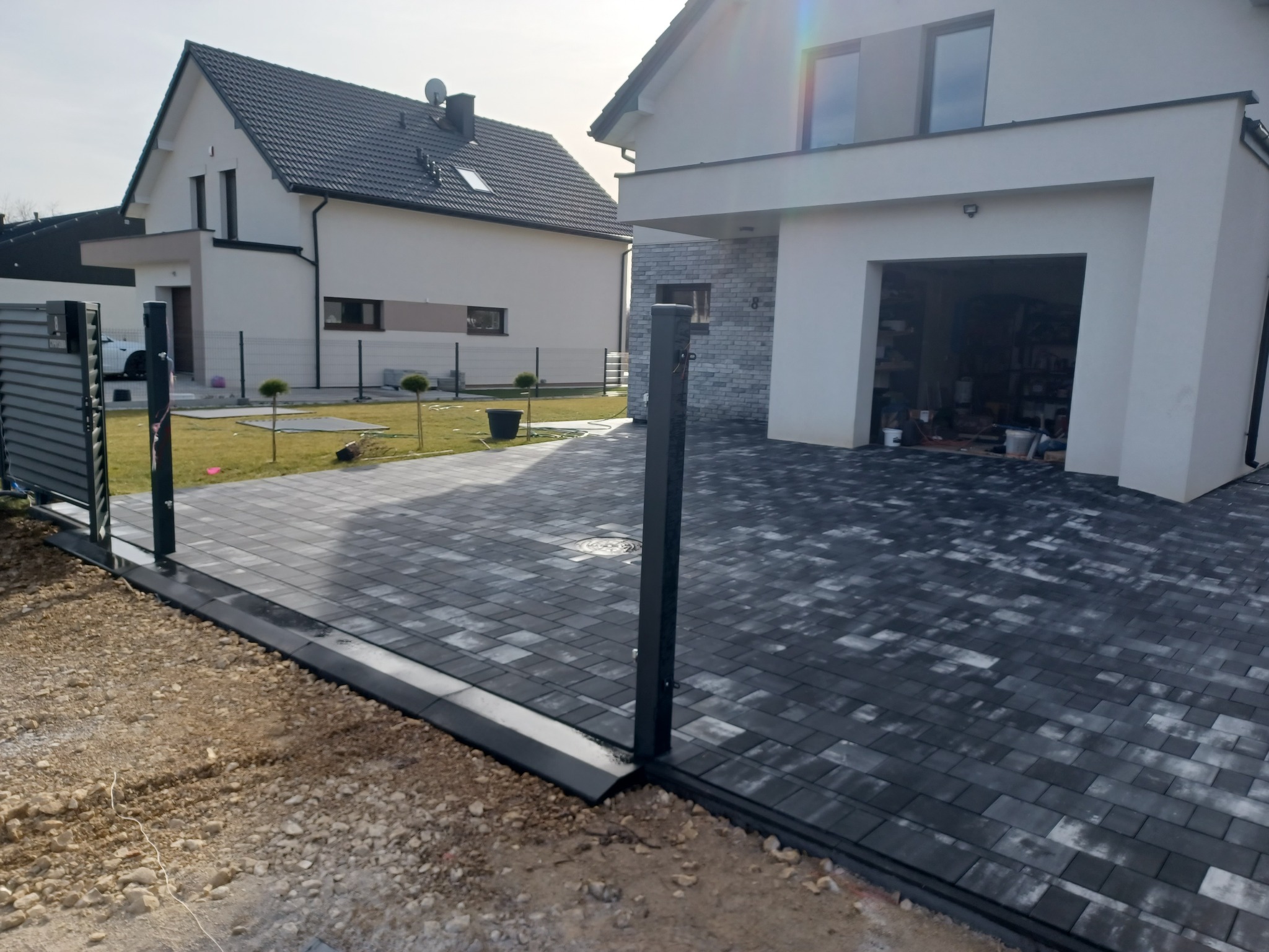 Ułożona, szara kostka brukowa na podjeździe do nowoczesnego domu z garażem, widoczne elementy bramy przesuwnej i przygotowane podłoże.