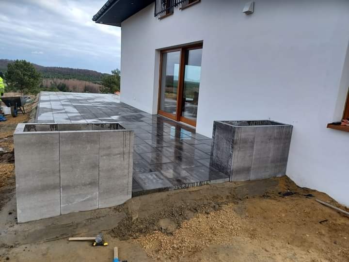 Taraz z kostki brukowej w trakcie budowy, z widocznymi donicami z betonowych elementów i narzędziami budowlanymi na ziemi.