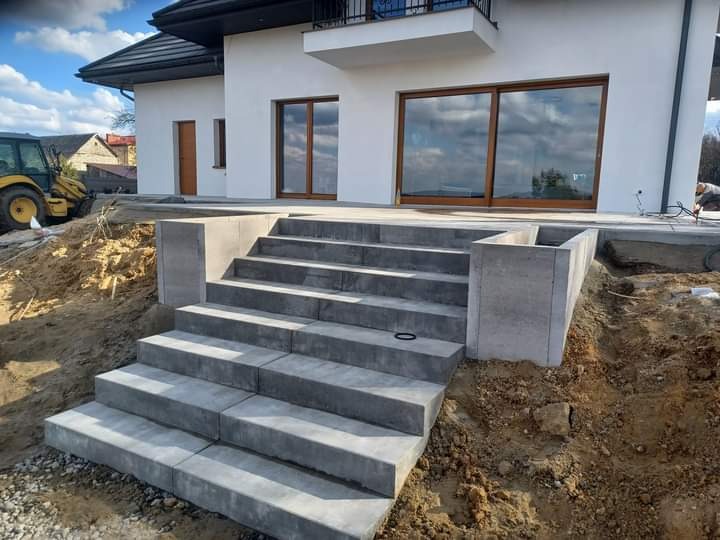 Schody zewnętrzne z szarej kostki brukowej prowadzące do nowoczesnego domu z dużymi oknami przesuwnymi, widoczny wykop i sprzęt budowlany w tle.