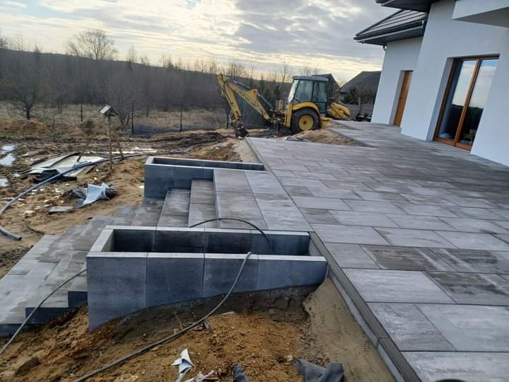 Układanie szarej kostki brukowej na tarasie przy nowoczesnym domu, z widocznym na tyłach żółtym koparko-ładowarką i przygotowanym terenem pod nasadzenia.