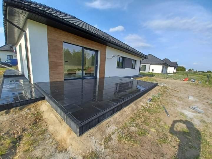 Nowoczesny taras z ciemnej kostki, przylegający do białego domu z dużym przesuwnym oknem, widoczny fragment ogrodu i inne domy w tle, świeżo ułożona kostka.