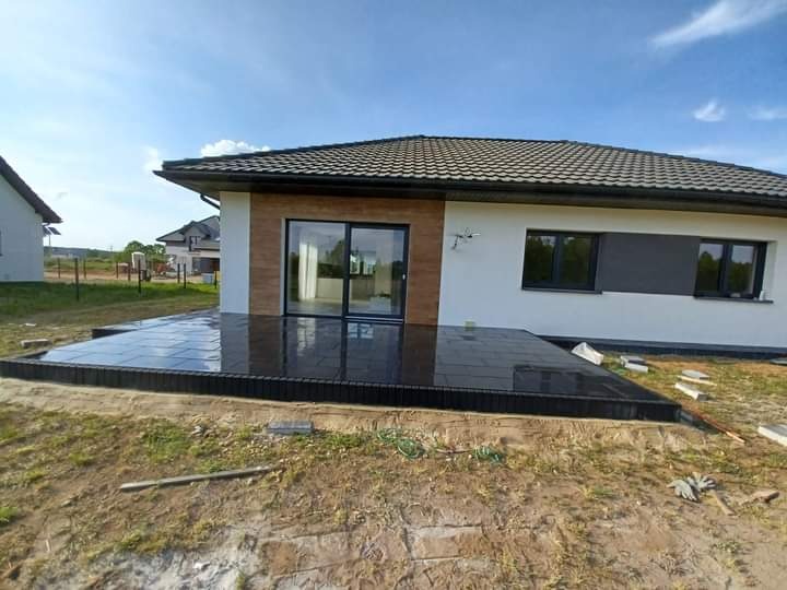 Nowoczesny taras z ciemnej, błyszczącej kostki przy białym domu z dużymi oknami przesuwnymi, widok na ogród w słoneczny dzień.