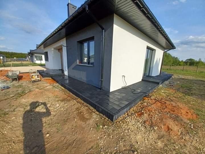 Świeżo ułożona, ciemnoszara kostka brukowa wokół nowoczesnego domu z szarą elewacją, widoczna ubijarka wibracyjna i cień fotografa.