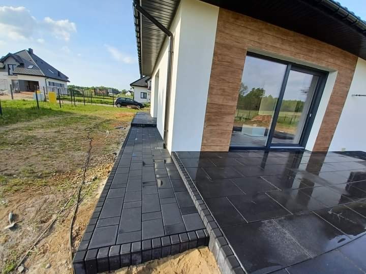 Wykończony taras i ścieżka z ciemnej kostki brukowej przy nowoczesnym domu jednorodzinnym, widoczne refleksy światła na powierzchni, w tle trawnik i drugi budynek.