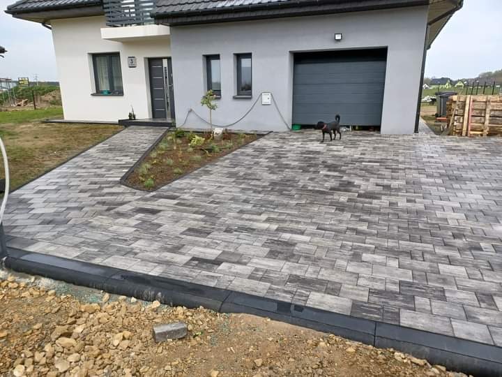 Ułożona kostka brukowa w odcieniach szarości prowadząca do domu z garażem, widoczny czarny pies na kostce i świeżo zasadzone rośliny wzdłuż ścieżki.
