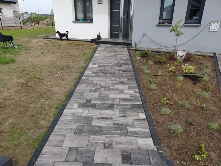 Ułożona ścieżka z kostki brukowej w odcieniach szarości prowadząca do wejścia domu z małym czarnym psem w tle i rabatą kwiatową po prawej stronie.