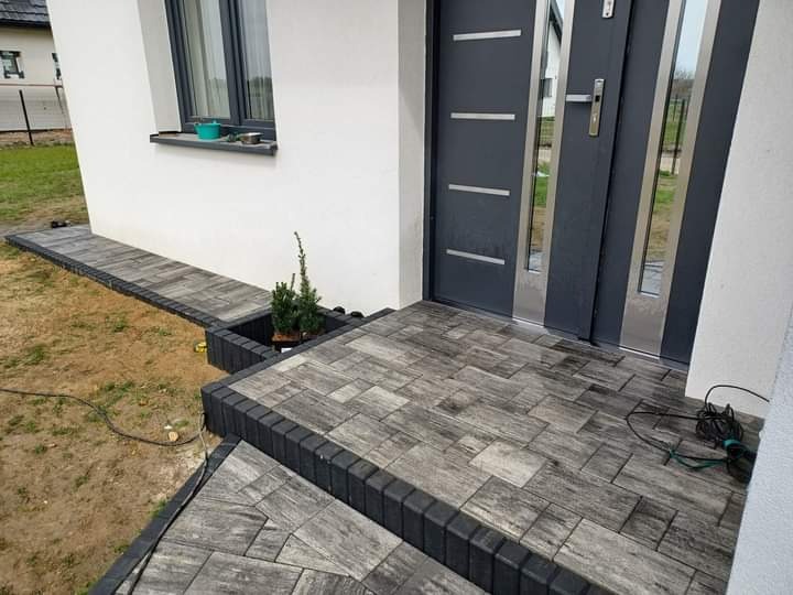 Nowoczesne wejście do domu z szarej kostki brukowej ułożonej w prostokątny wzór, obrzeża z ciemnej kostki, mała rabata z iglakiem przy schodach, szare drzwi wejściowe z przeszkleniami.