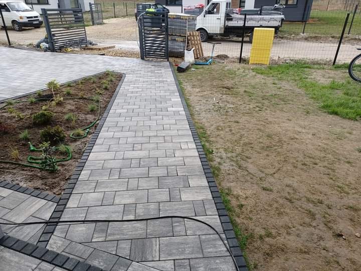Ułożona ścieżka z szarej kostki brukowej z ciemnym obramowaniem, prowadząca od schodów do furtki, obok rabata z młodymi roślinami i trawnik.