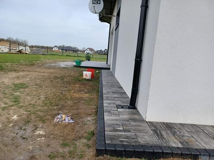 Ułożona ścieżka z szarej kostki brukowej przy białej ścianie budynku z czarną rynną spustową. W tle widoczny teren budowy i domy.