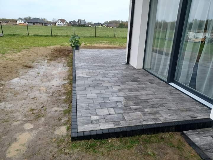 Wykończony taras z szarej kostki brukowej ułożonej w regularny wzór, obrzeże z ciemnej kostki, widok na trawnik i domy w tle.