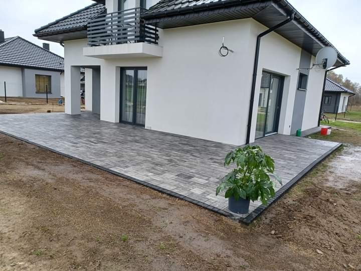 Nowoczesny taras z szarej kostki brukowej ułożonej przy białym domu z balkonem, donica z zieloną rośliną w rogu tarasu.