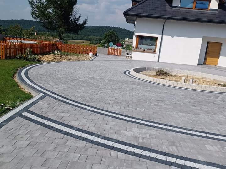 Szeroki podjazd z szarej kostki brukowej ułożonej w regularny wzór, z dekoracyjnymi pasami z jasnej kostki i obrzeżem z ciemnej kostki, prowadzący do białego domu z brązowymi drzwiami, widok...