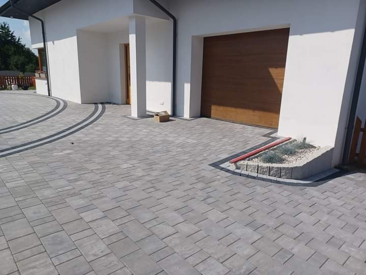 Szara kostka brukowa ułożona przed nowoczesnym domem z brązową bramą garażową, z dekoracyjnym obramowaniem z ciemniejszej kostki i małą rabatą z roślinami.