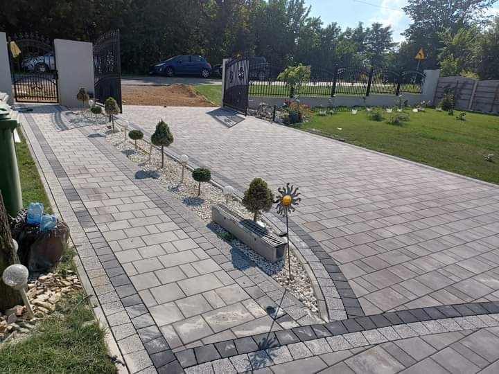 Ułożona szara kostka brukowa na podjeździe z dekoracyjnym obramowaniem, widoczny ogród z metalową bramą i fragment domu w tle.